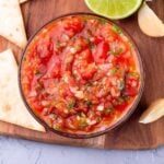 A bowl of simple, authentic Mexican restaurant-style salsa with diced tomatoes, onions, cilantro, and lime juice sits surrounded by tortilla chips, garlic cloves, and a sliced lime on a wooden board.