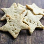 Star-shaped brown butter cookies with faint imprints on top are arranged on a wooden surface. The cookies appear to be homemade with a light golden color, infused with bourbon notes that convey a sense of warmth and simplicity.