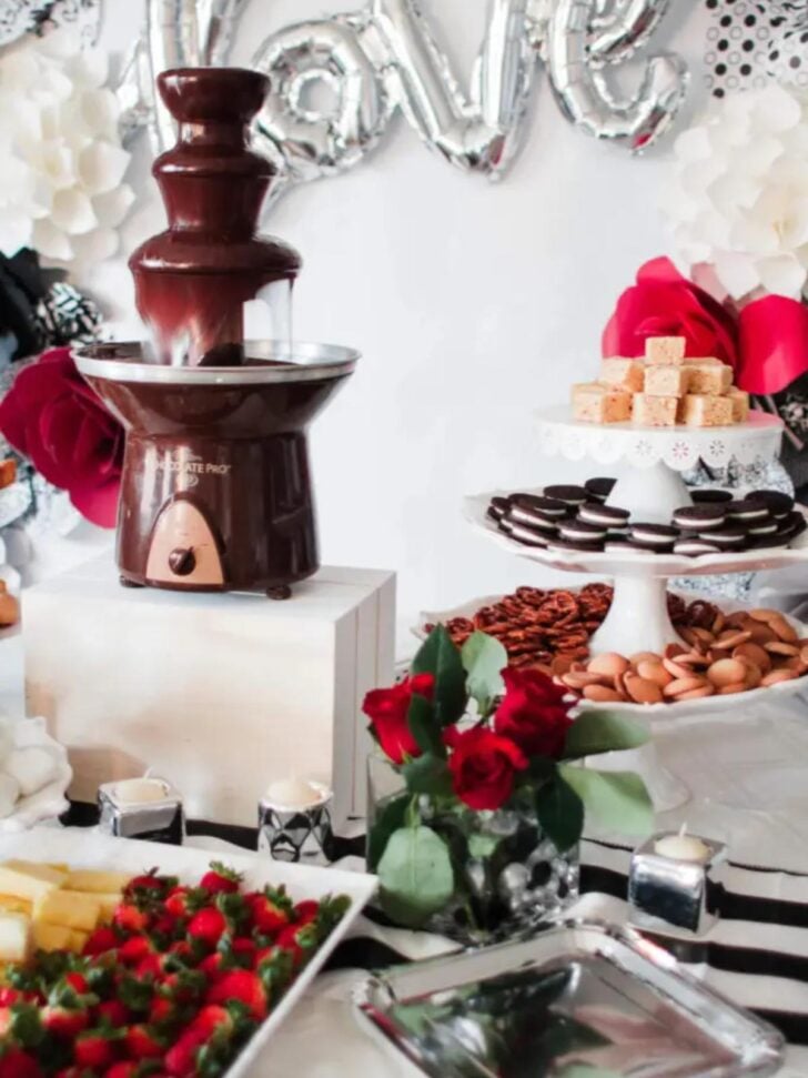 A chocolate fountain on a table surrounded by trays of fruit, strawberries, cookies, pretzels, and small cakes—delicious food bar ideas. Red roses, candles, and a silver "love" balloon decorate the festive display.