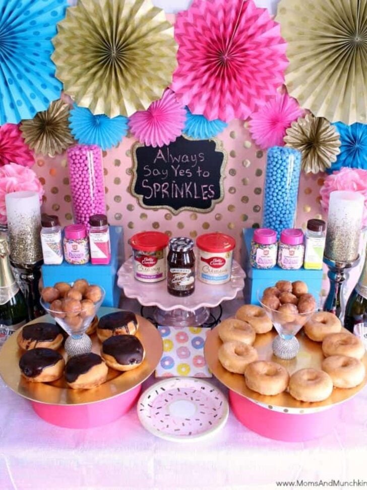 A colorful dessert table, perfect for snack bar ideas, features donuts, donut holes, jars of sprinkles, frosting, and toppings. Pink, gold, and blue paper fans decorate the background. A chalkboard sign reads, “Always Say Yes to Sprinkles.”.