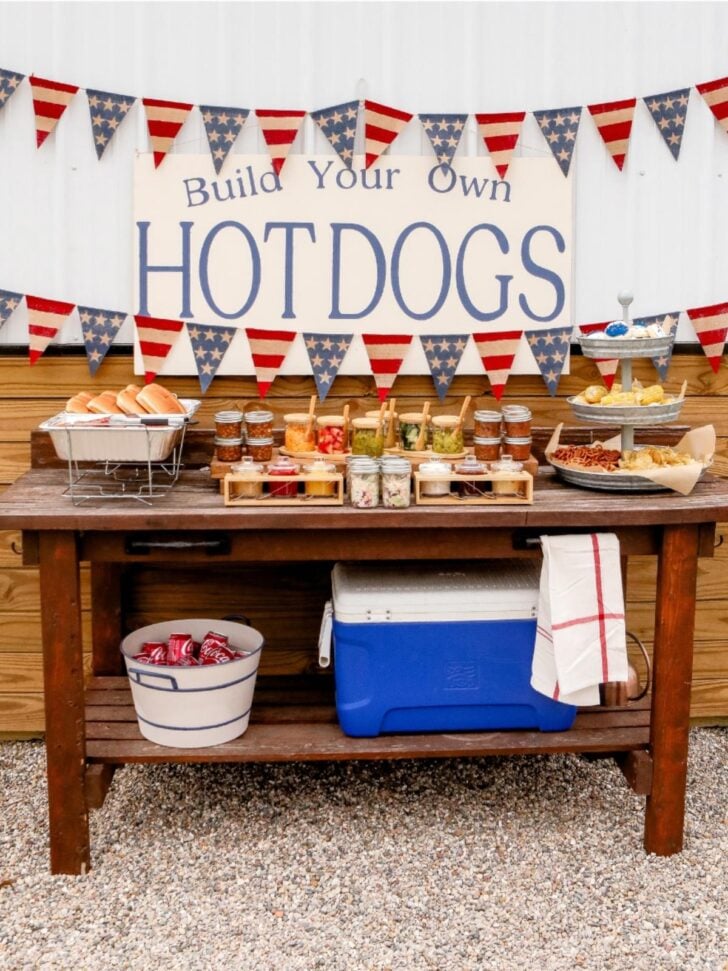 A rustic outdoor table is set up as a “Build Your Own Hot Dogs” station with buns, various toppings in jars, chips, and a cooler—perfect for those looking for festive food bar ideas. Red, white, and blue bunting decorates the background.