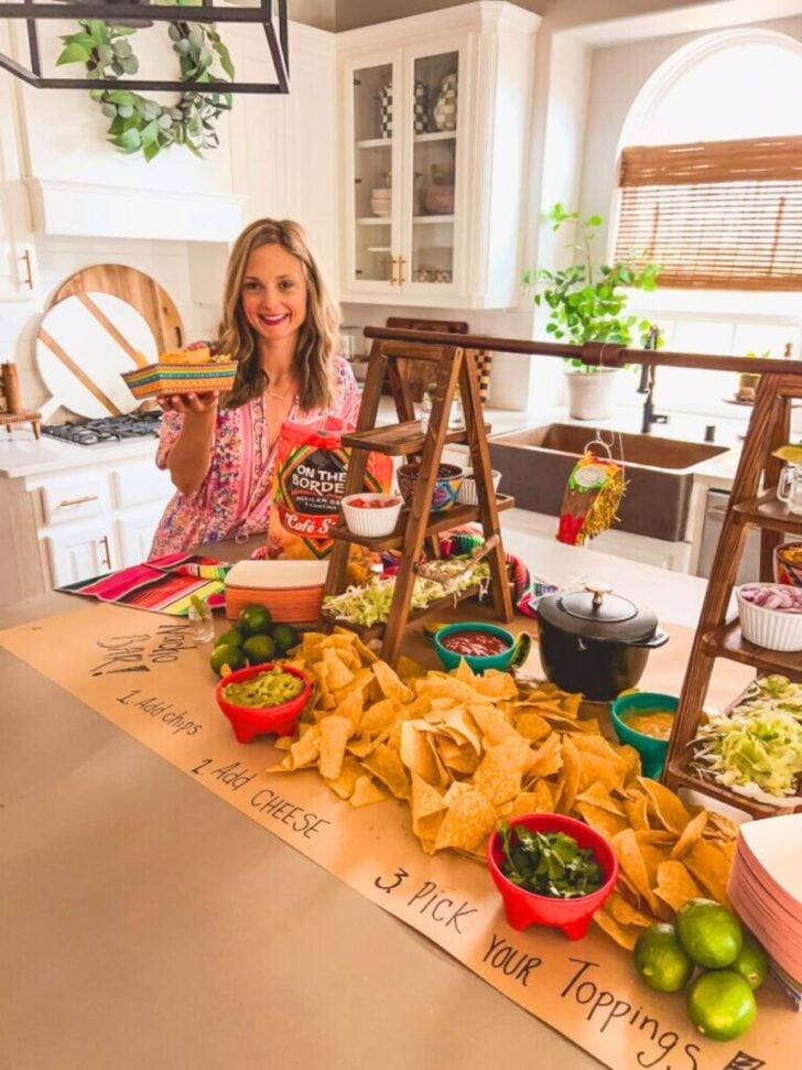 A woman stands in a bright kitchen holding a taco, smiling behind a counter filled with bowls of taco toppings, chips, limes, and salsa—a perfect scene for those looking for creative food bar ideas.