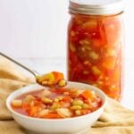 A bowl of canned vegetable soup with various vegetables is placed on a light-colored cloth beside a jar filled with the same soup. A spoon is lifting a portion from the bowl, showcasing chunks of vegetables in a tomato-based broth.
