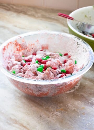 A bowl of checks covered in white chocolate that has been dyed red and sprinkled powdered sugar on with a few red and green M&Ms tossed in