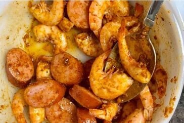 A bowl filled with cooked shrimp and sliced sausage is coated in a spicy, seasoned sauce reminiscent of a boiling crab sauce recipe. A metal spoon rests in the bowl, partially submerged in the flavorful mix. The dish is rich in color, showcasing a glossy texture.