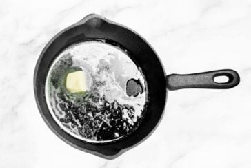 A black cast iron skillet on a white surface holds a melting pat of butter, surrounded by bubbling liquid reminiscent of a savory boiling crab sauce recipe.