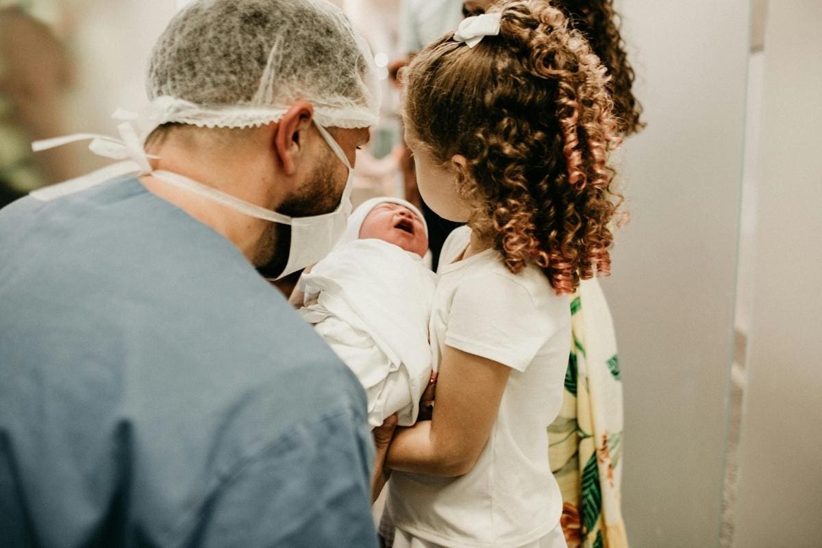 A man in medical scrubs and a hairnet looks at a curly-haired girl holding a newborn wrapped in a white blanket. The girl gazes gently at the baby in a hospital setting, inspiring thoughts of how to start a meal train for new parents.