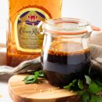 A glass jar filled with a dark liquid and floating garlic pieces sits on a wooden board, surrounded by green herbs. Behind the jar, there is an amber-colored bottle of Crown Royal whiskey, hinting at a rich whiskey marinade. A cloth napkin is draped around the items.