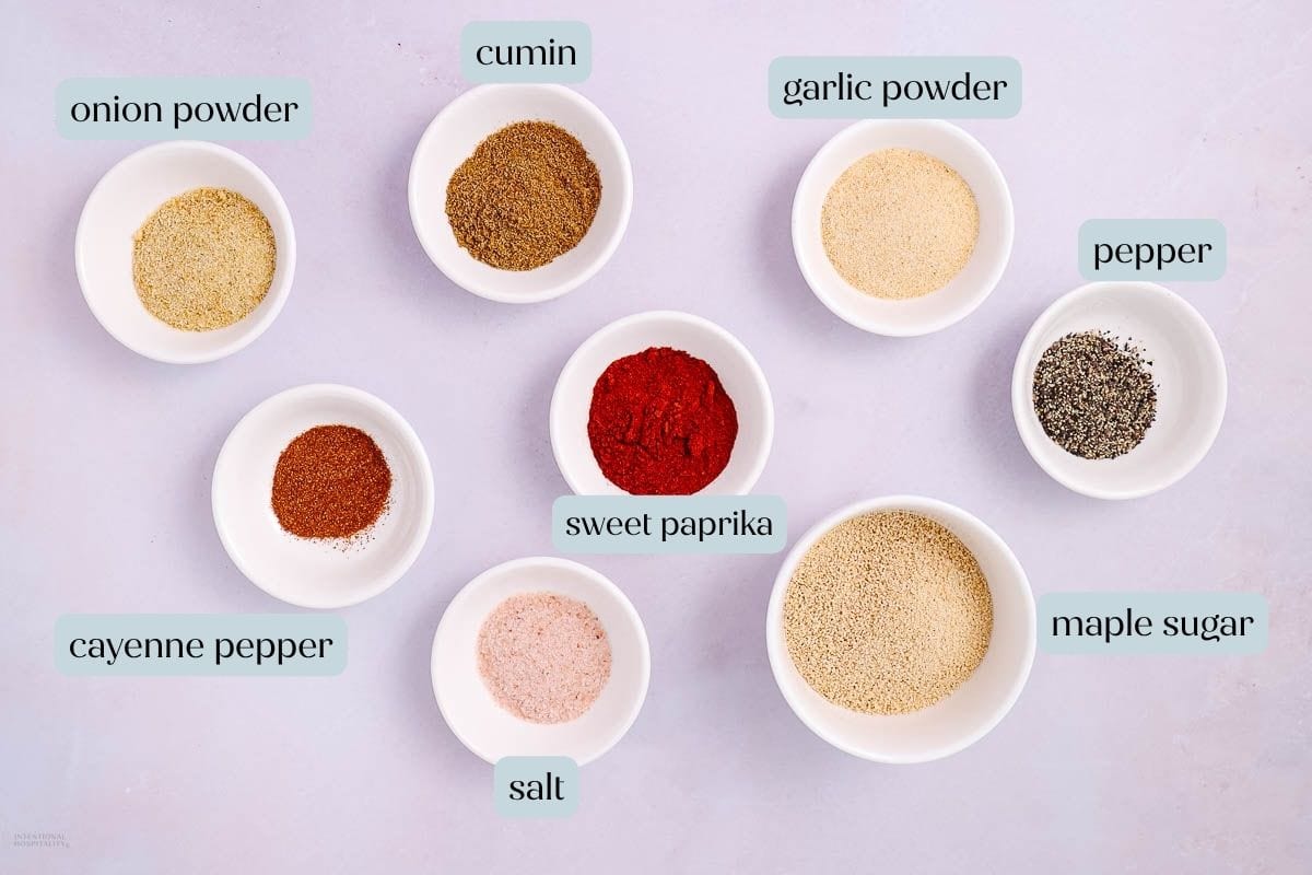 Eight small bowls with various spices and seasonings, each labeled: onion powder, cumin, garlic powder, pepper, maple sugar, salt, cayenne pepper, and sweet paprika—perfect ingredients for a sweet BBQ Dry Rub—are arranged on a light surface.