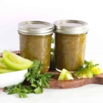 two jar of green tomato salsa with fresh green tomatoes by them and cilantro