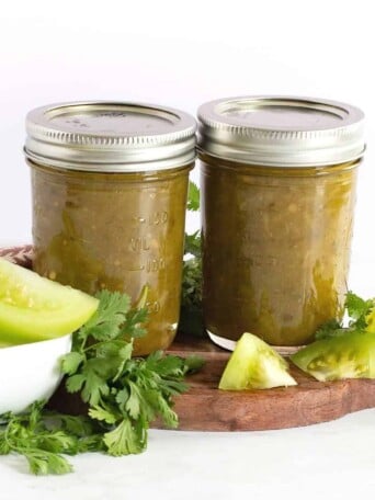 two jar of green tomato salsa with fresh green tomatoes by them and cilantro
