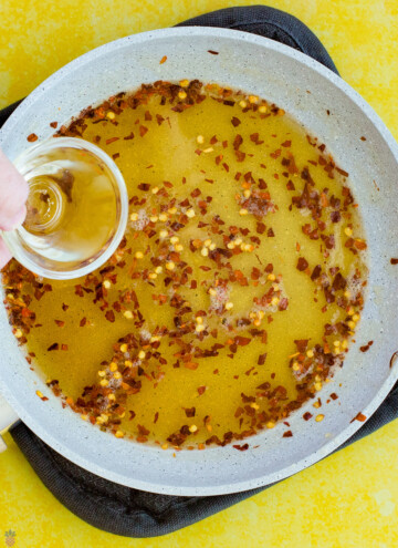 honey and chili flakes and seeds in a pan with vinegar being poured in.