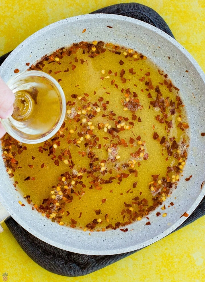 honey and chili flakes and seeds in a pan with vinegar being poured in.