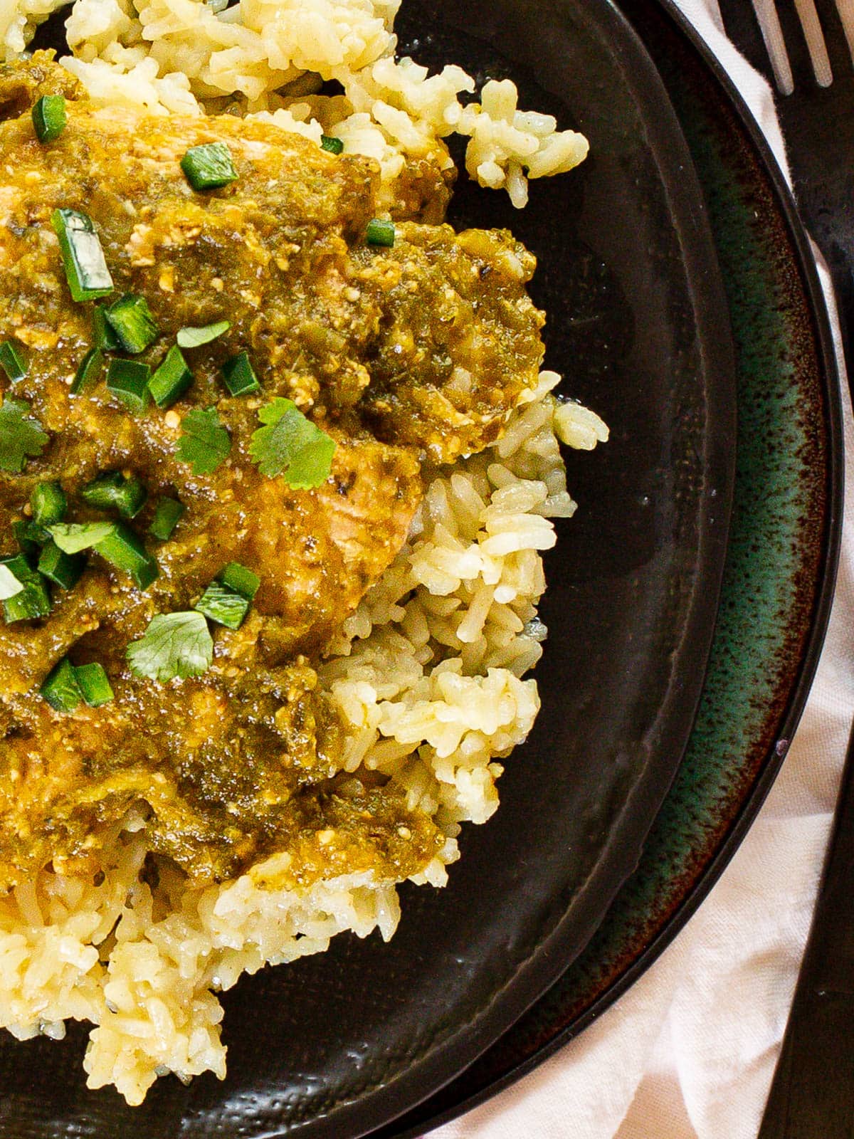 A close-up of a plate with yellow rice topped with a vibrant green sauce from a pollo verde recipe and garnished with chopped green onions. A fork is placed beside the plate.