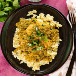 A black plate filled with white rice topped with chicken in a green sauce, garnished with chopped green herbs. Nearby are a fork, knife, napkin, and fresh cilantro—perfect for serving your favorite pollo verde recipe.