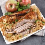 Oven baked pork and sauerkraut on a white platter, with seasoned pork roast pulled into thick pieces over sauerkraut and soft apple chunks.