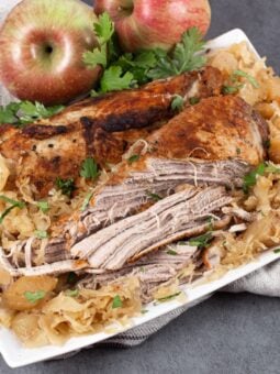 Oven baked pork and sauerkraut on a white platter, with seasoned pork roast pulled into thick pieces over sauerkraut and soft apple chunks.
