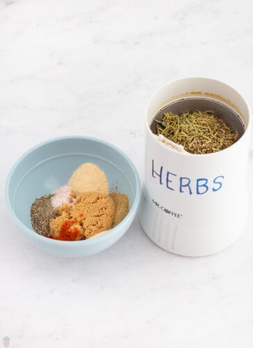 whole herbs, such as oregano and rosemary in a spice grinder with salt, pepper, brown sugar and garlic powder in a small bowl.