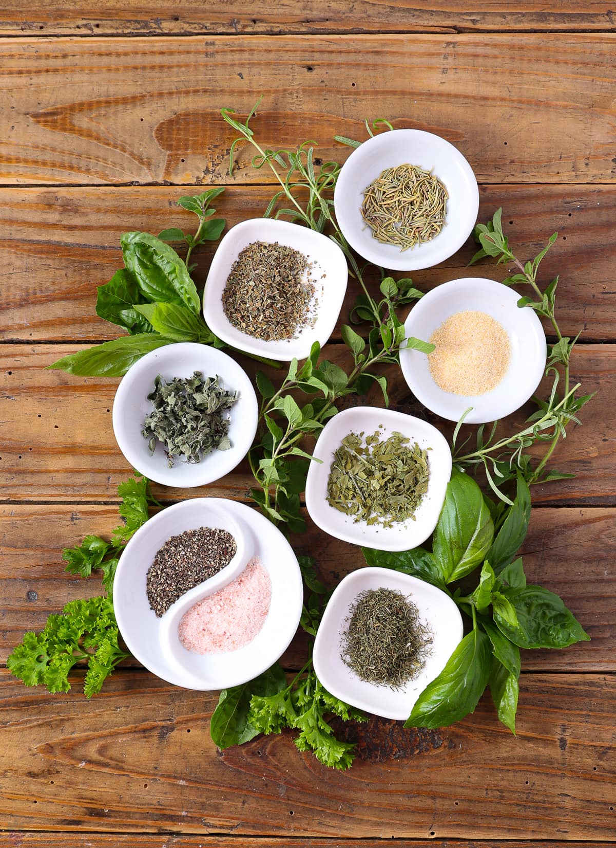 A display of nine white bowls filled with various herbs and spices arranged on a wooden surface. Fresh green herbs, including basil, are scattered around the bowls. The spices resemble those in a Texas Roadhouse chicken recipe, adding a vibrant and earthy feel to the scene.