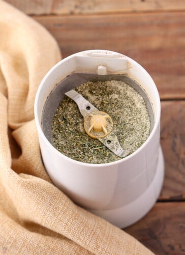 A white spice grinder filled with dried herbs and spices sits on a wooden surface. The grinder's top is open, revealing its metal blades surrounded by the ground mixture perfect for a Texas Roadhouse chicken recipe. A beige cloth is draped beside the grinder.
