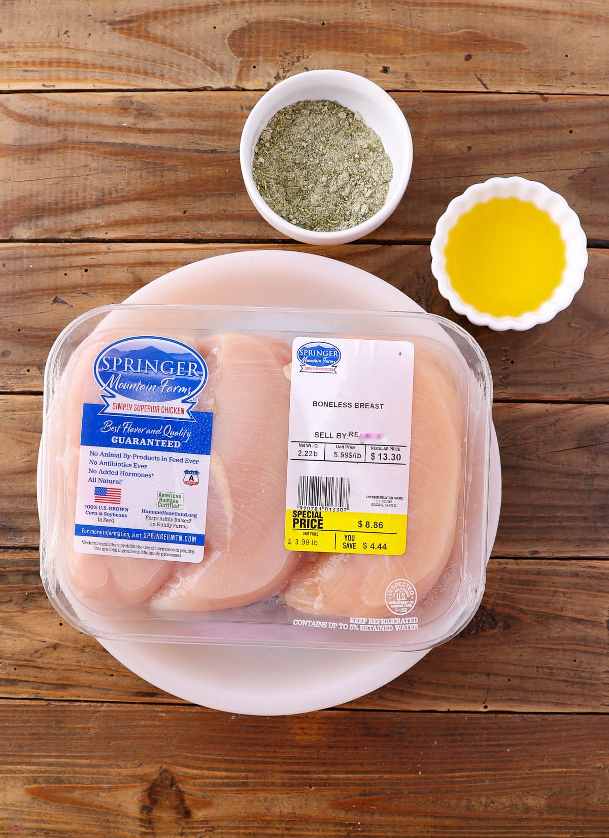 A package of boneless chicken breast labeled "Springer Mountain Farms" sits on a white plate. Next to it, there's a small bowl of green herb mixture and a small white bowl containing yellow olive oil, perfect for crafting a Texas Roadhouse chicken recipe. The items are placed on a rustic wooden surface.