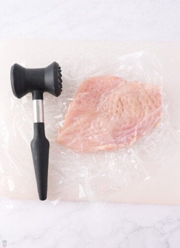 A raw chicken breast, partially covered with plastic wrap, is placed on a cutting board. A meat tenderizer, featuring a black handle and a dual head with one flat and one textured side, is positioned next to the chicken breast—perfect prep for your favorite Texas Roadhouse chicken recipe.