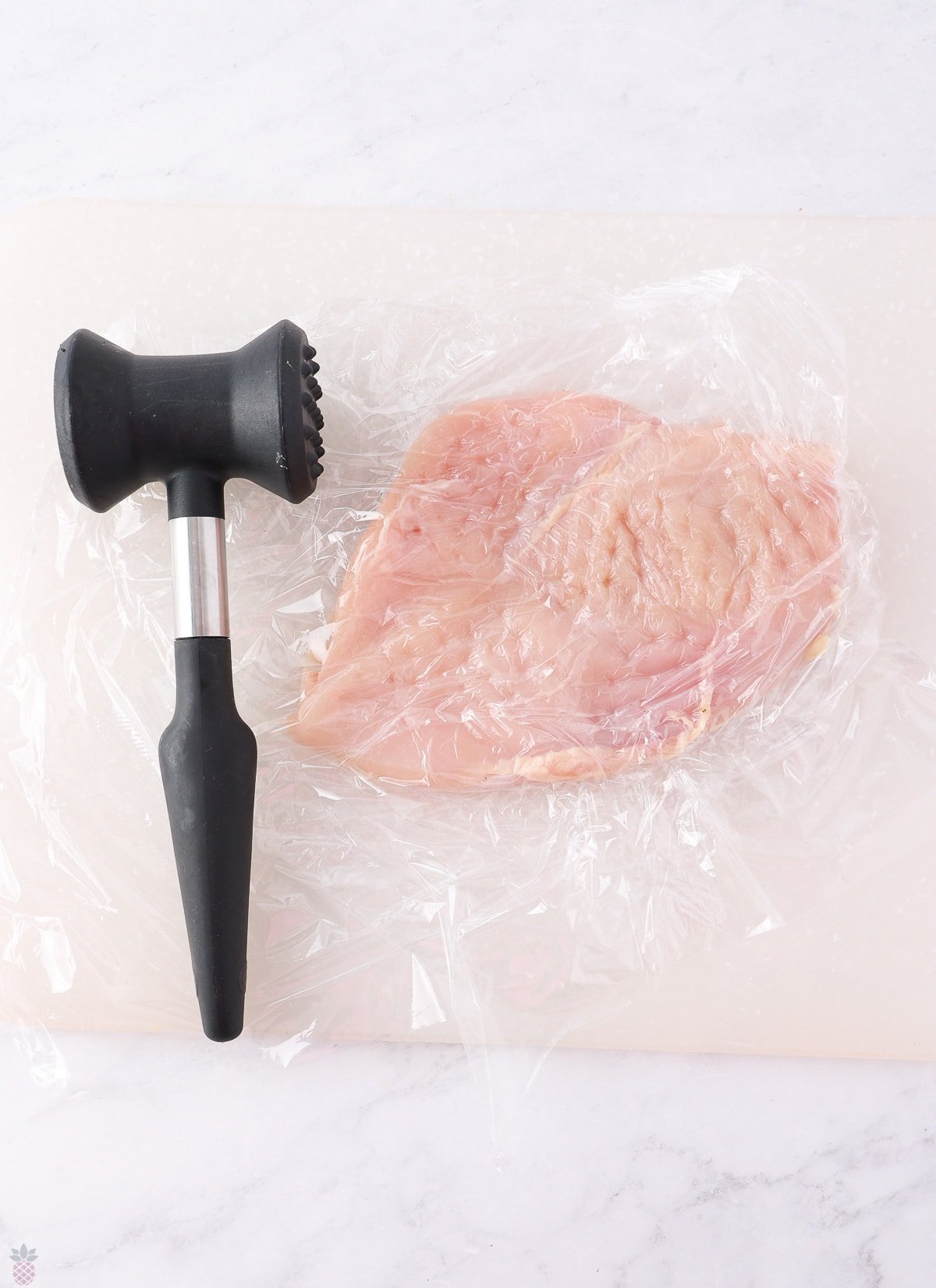 A raw chicken breast, partially covered with plastic wrap, is placed on a cutting board. A meat tenderizer, featuring a black handle and a dual head with one flat and one textured side, is positioned next to the chicken breast—perfect prep for your favorite Texas Roadhouse chicken recipe.