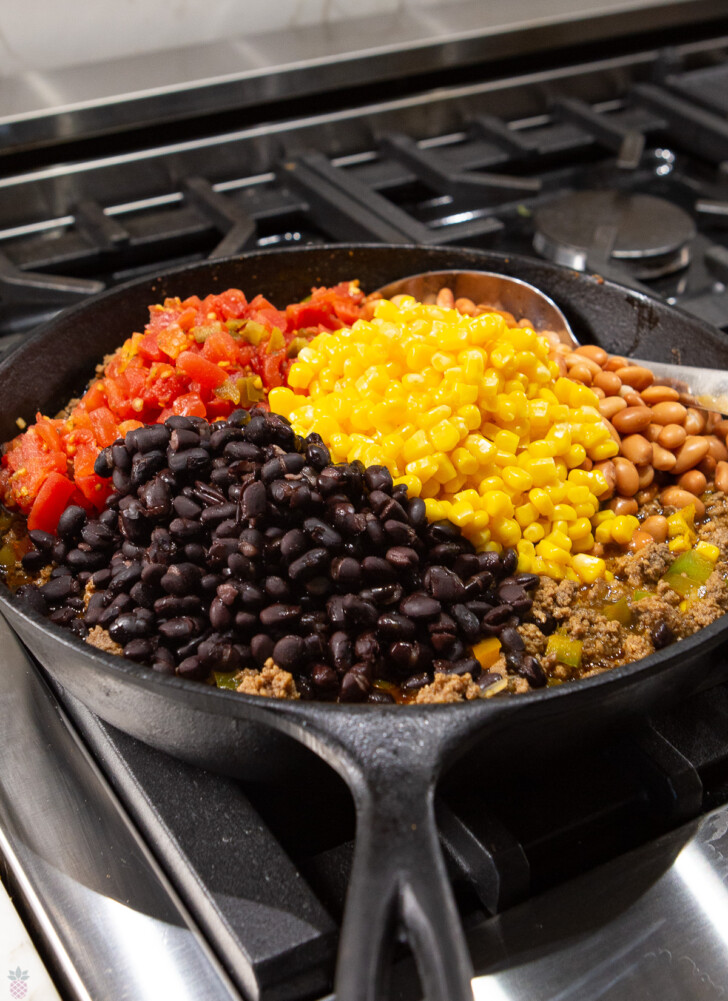 slow cooker mexican casserole ground beef in black skillet with corn, black beans and tomatoes being stired in.