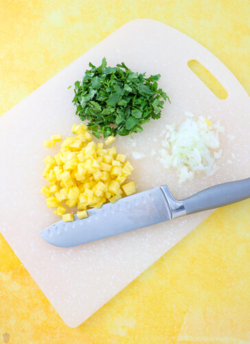 Chopped onions pineapple and cilantro on a cutting board with a knife near them