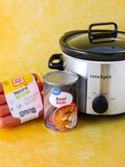 A package of turkey franks, a can of beef broth, and a Crock-Pot slow cooker are arranged on a yellow surface with a matching yellow background.