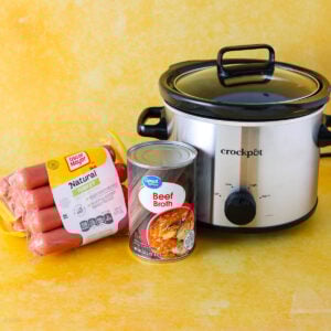 A package of turkey franks, a can of beef broth, and a Crock-Pot slow cooker are arranged on a yellow surface with a matching yellow background.