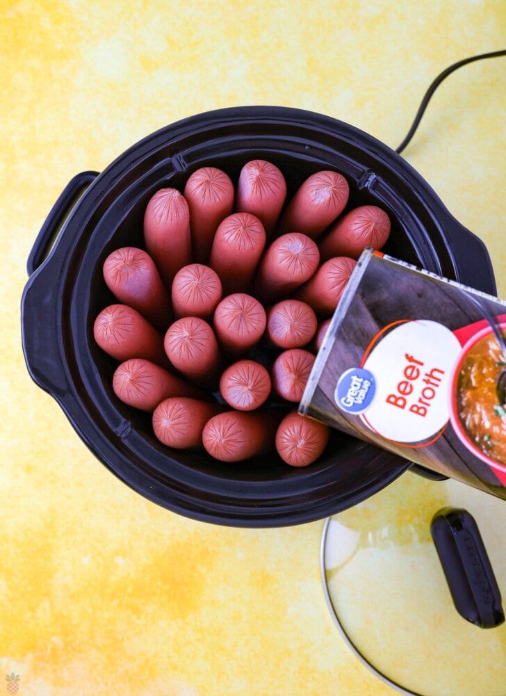 hot dogs in a slow cooker with a can of beef broth being poured over them.
