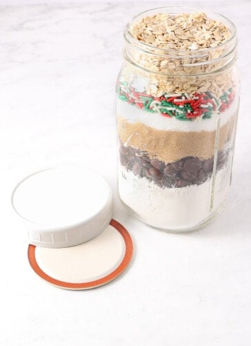A layered mason jar with cookie mix and a Ball canning jar lid and plastic lid sitting next to it ready to seal up the jar