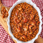 A big pot of Boston baked beans that are Saucy with big chunks of bacon and a spoon with beans on it ready to serve