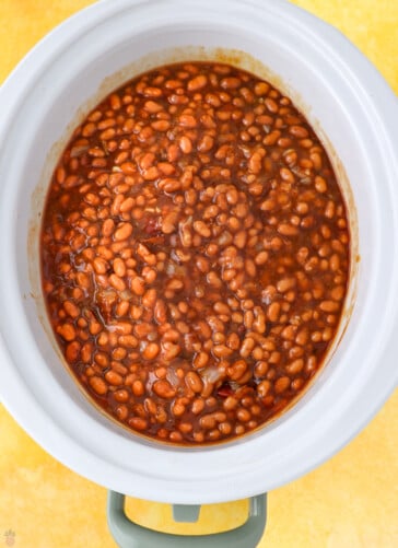 A white slow cooker filled with a delicious baked bean recipe in crock pot style, featuring tender beans in a thick, reddish-brown sauce, set on a yellow surface.