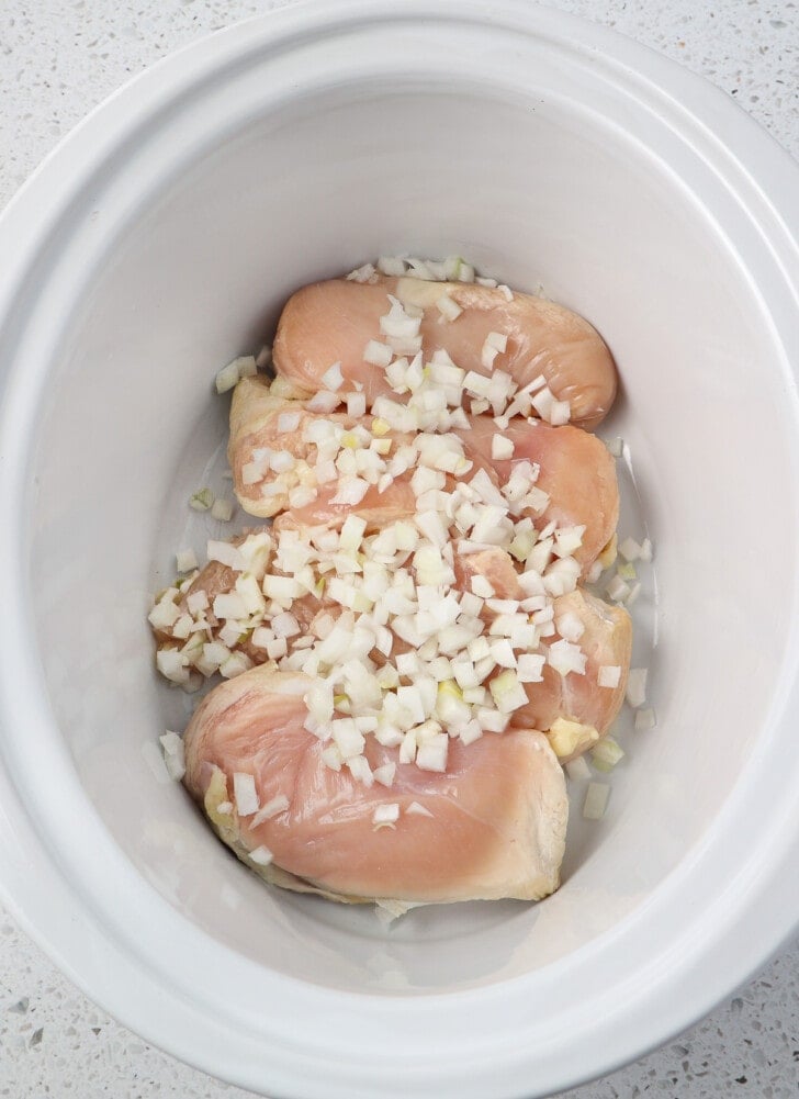 Raw chicken breast in a crock pot with diced onions sprinkled on top