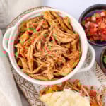 A bowl of Crock-Pot chicken tacos sitting next to a taco shell stuffed with meat cheese and sour cream and a Sprinkle of salsa