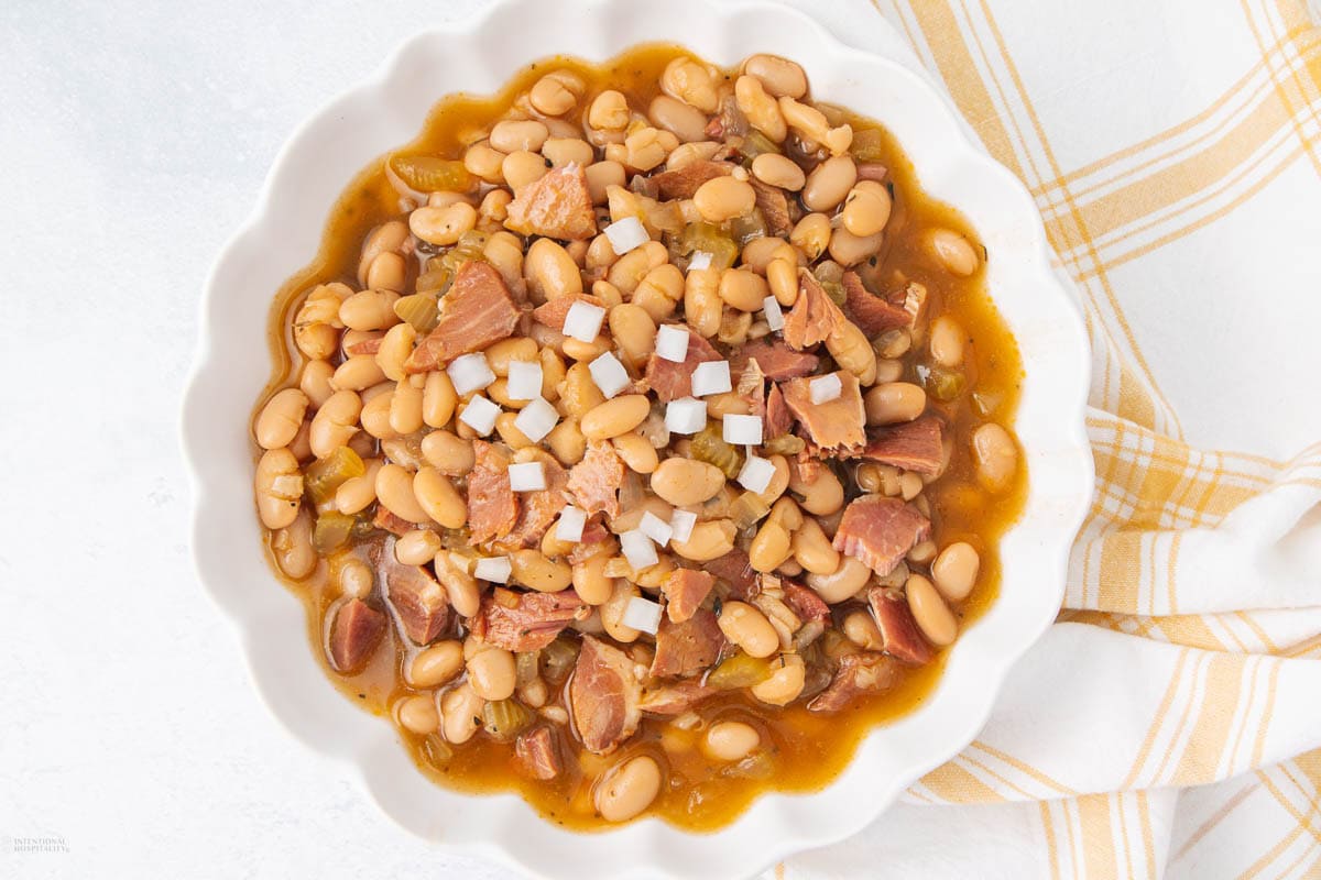 A white bowl filled with baked navy beans, pieces of bacon or ham, and topped with diced onions, placed next to a yellow and white checkered cloth on a white surface.