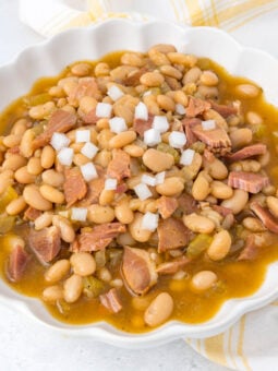 A white bowl filled with ham and navy bean soup, topped with diced white onions. The soup contains navy beans, chunks of ham, and vegetables in a golden broth. A yellow and white napkin is in the background.