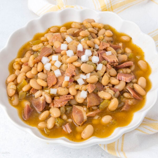 A white bowl filled with ham and navy bean soup, topped with diced white onions. The soup contains navy beans, chunks of ham, and vegetables in a golden broth. A yellow and white napkin is in the background.
