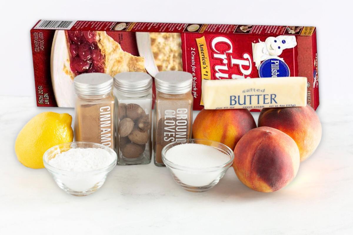 A box of pie crust mix, a stick of butter, fresh peaches, a lemon, jars of cinnamon, nutmeg, and ground cloves, plus bowls of sugar and powdered sugar—everything you need for an old fashioned peach cobbler on a white surface.