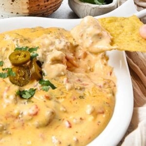 A chip is dipped into a creamy Rotel sausage dip with visible chunks of tomato and ground meat, topped with sliced jalapeños and chopped cilantro, served in a white dish.