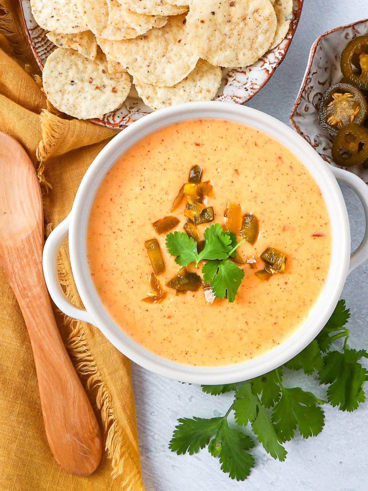 A bowl of creamy queso dip garnished with cilantro and diced jalapeños, surrounded by tortilla chips, a small bowl of sliced jalapeños, a wooden spoon, and fresh cilantro on a yellow napkin.