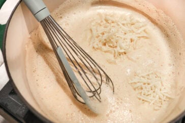 A metal whisk stirs shredded cheese into a pot of foamy liquid, possibly creating a creamy queso dip or sauce, on a stovetop.