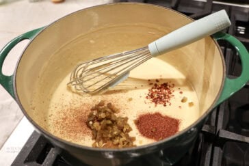 A whisk rests in a pot of creamy queso dip on the stove, with piles of chili powder, diced green chilies, and red pepper flakes on top, ready to be mixed in.