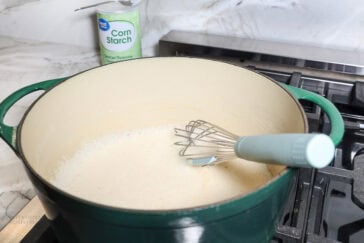A whisk rests in a large green pot on a stove, mixing a white powdery substance for queso dip. In the background, there is an open container of corn starch on a marble countertop.