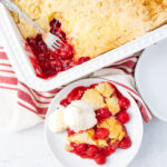 A white ceramic baking dish filled with cherry cobbler, partially scooped out, is next to a white plate holding a serving of cherry cobbler topped with a scoop of vanilla ice cream. A red striped towel is placed under the plate on a white surface.