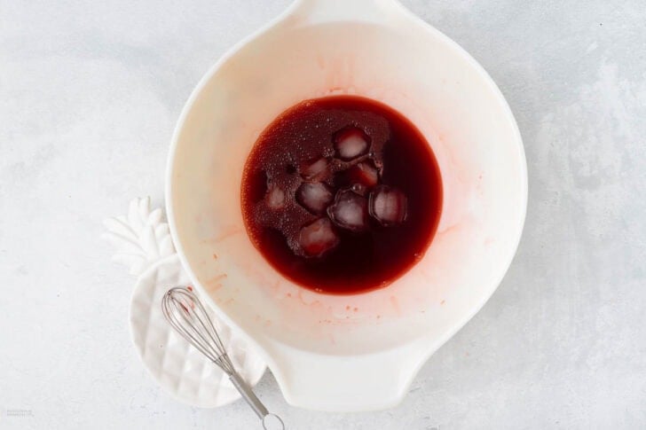 A white bowl containing a red liquid with ice cubes sits on a light gray surface. Next to the bowl is a small white plate with a miniature whisk resting on it. The overall background and objects are minimalistic.