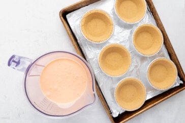 A baking tray lined with foil holds six small, empty pie crusts. Next to the tray is a mixing pitcher filled with a light orange liquid mixture. The scene is set against a light-colored countertop.