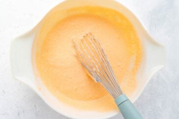 A white mixing bowl contains a frothy, orange-colored batter. A whisk with a light blue handle rests inside the bowl, indicating that the batter has been recently mixed. The bowl is placed on a light gray, textured surface.